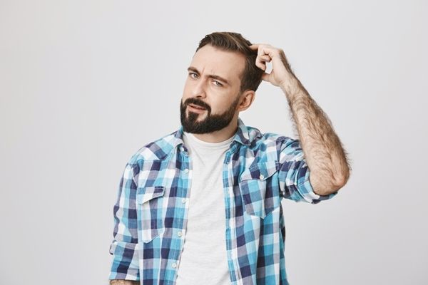 Edad para hacer injerto de cabello - hombre blanco con camiseta blanca y camisa a cuadros rascándose la cabeza de forma pensativa