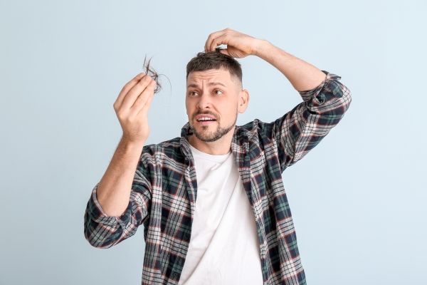Edad para hacer injerto de cabello - hombre joven con camiseta blanca y camisa de manga larga a cuadros mirando atónito el mechón de pelo que tiene en la mano