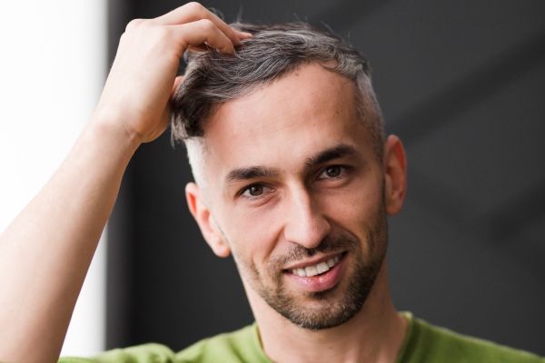 hombre joven sonriente con el cabello corto y con canas acariciando su cabello con la mano 