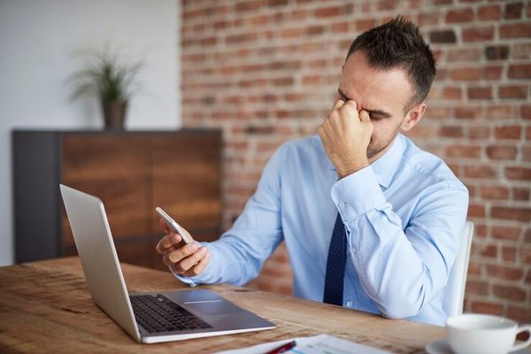 alopecia nerviosa - hombre sentado en la oficina delante de la computadora, sujetando el móvil en la mano derecha y la mano izquierda en los ojos, con signos de alopecia en las entradas del cabello
