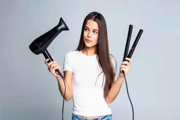 mujer con camiseta blanca y jeans mirando un secador y una plancha para el pelo decidiendo cual va a usar