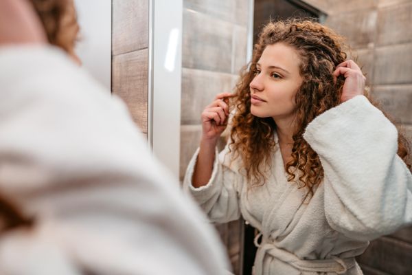 aceite de coco en el pelo - mujer joven en el baño con un albornoz revisando su cabello