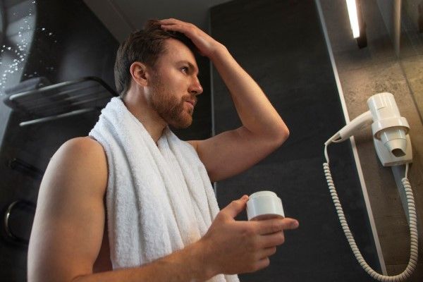 Hombre recién salido de la ducha se mira al espejo mientras aplica un producto capilar especial para potenciar el brillo y la hidratación de su melena 