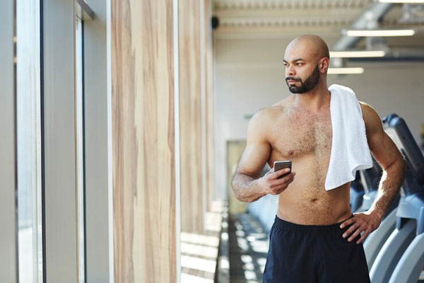 Testosterona y caída del cabello - Hombre calvo, musculoso y con barba mira por la ventana mientras se toma un break en el gimnasio 