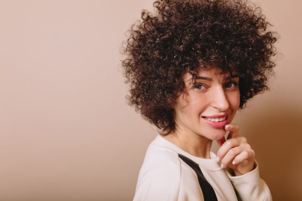 densidad del cabello rizado - Amable mujer linda con rizos y maquillaje desnudo sonríe a la cámara y disfruta de buen humor
