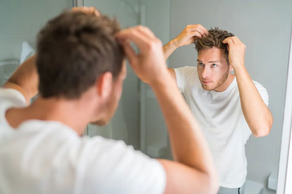 Alopecia en jóvenes - Joven de camisa blanca chequea el estado de su cabello frente al espejo tras aplicar un tratamiento capilar