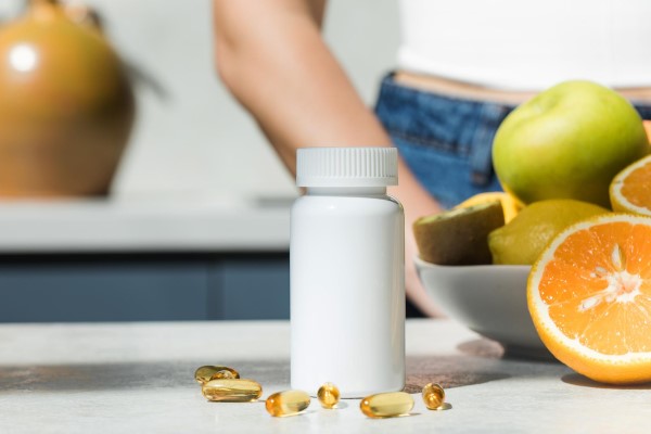  bote de capsulas sobre un mesa con un bol de fruta a su lado y una mujer detrás