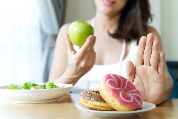 Mujer adulta con manzana verde en mano rechaza un plato de donas glaseadas para empezar a adoptar hábitos alimenticios más saludables