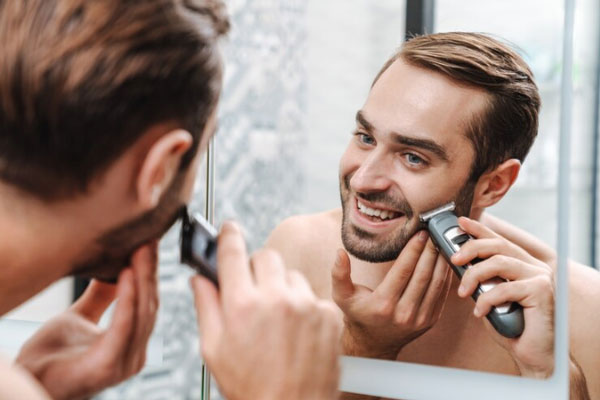 Cuidar la barba - Hombre de ojos azules sonríe mientras le da forma a su barba frente al espejo con una máquina de afeitar eléctrica 