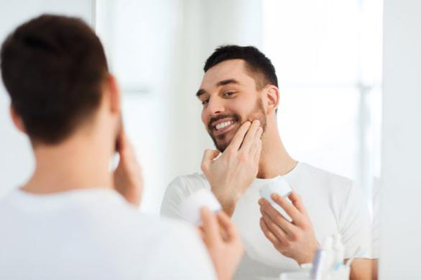 Cuidar la barba - Joven sonríe frente al espejo mientras aplica una crema hidratante en su rostro 