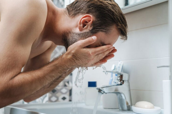 Cuidar la barba - Hombre se lava la cara con abundante agua en el lavamanos como parte de su rutina de limpieza facial 