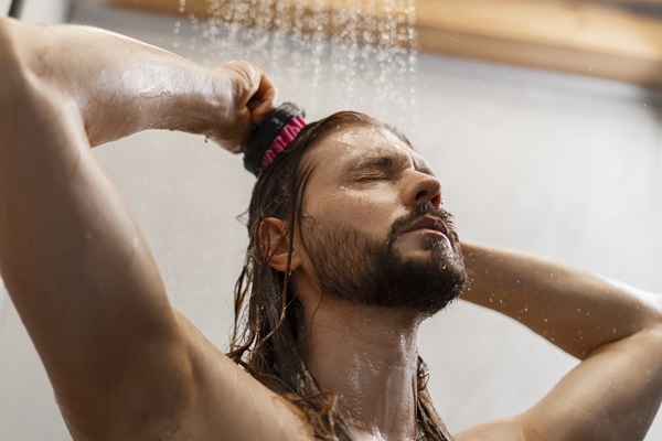 Caballero debajo de la ducha lava su cabello mientras da masajes al cuero cabelludo