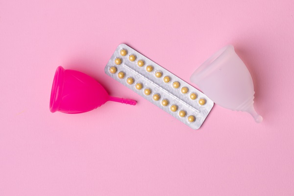 Espironolactona - Copa menstrual de color fucsia y blanca junto a pastillas anticonceptivas sobre una mesa rosada