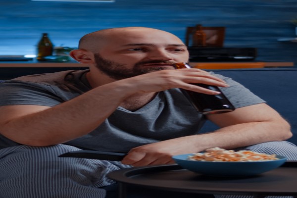 hombre con problemas graves de alopecia bebiendo cerveza sentado en un sofá con un bol de palomitas de maíz