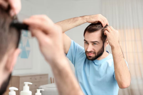 Hombre preocupado y con entradas en la cabeza peina su cabello frente al espejo 