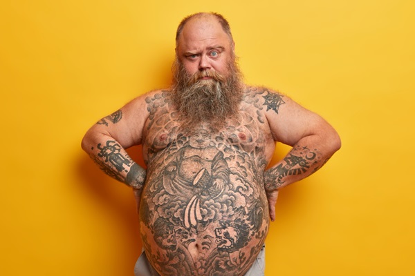 Calvicie y obesidad - Hombre con pérdida de cabello y tatuajes en el cuerpo mostrando su gran barriga a causa de la obesidad