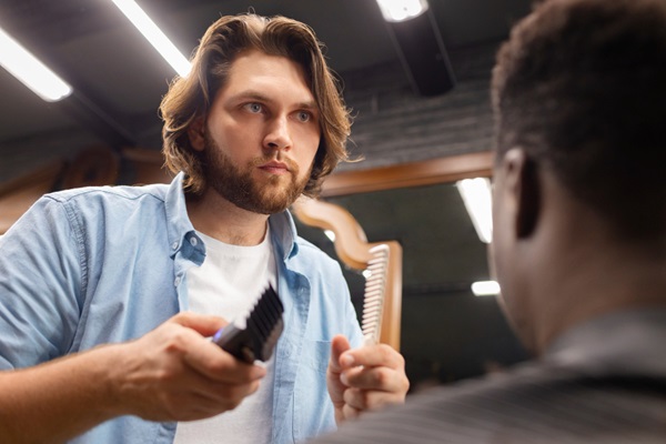 Estilista con el cabello largo en forma de libro abierto, sosteniendo una máquina de afeitar y un cepillo frente a un cliente