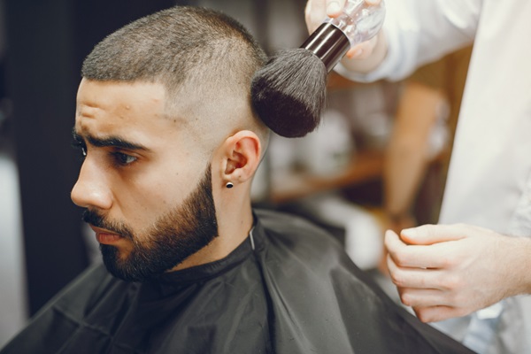 Cortes de pelo para hombres - Estilista usando una brocha para limpiar el corte buzz en el cabello de un cliente masculino