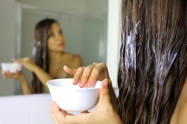 Mascarillas para hidratar el cabello - Mujer joven observándose en el espejo mientras se aplica una mascarilla hidratante para el cabello